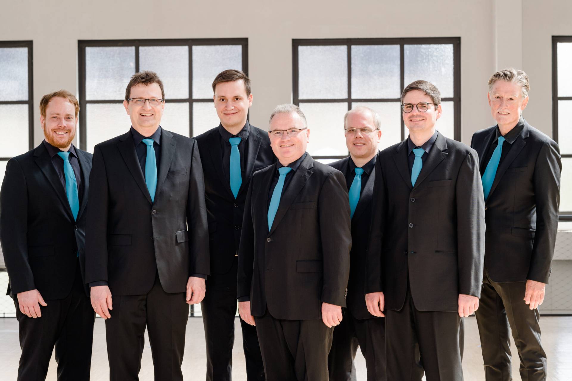 Sieben lächelnde Männer in schwarzen Anzügen und blauen Krawatten vor einer hellen Fensterfront
