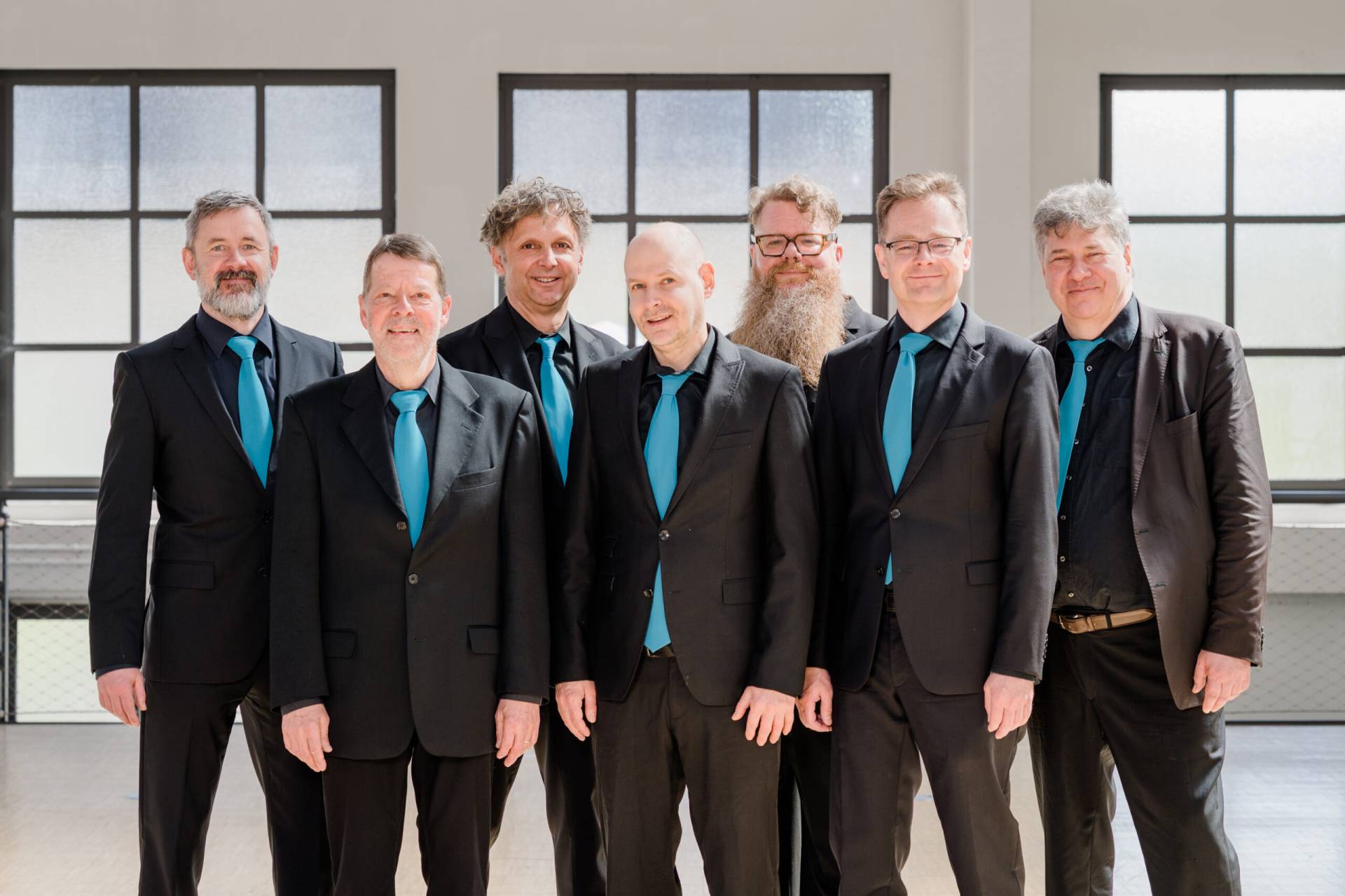 Sieben lächelnde Männer in schwarzen Anzügen und blauen Krawatten vor einer hellen Fensterfront