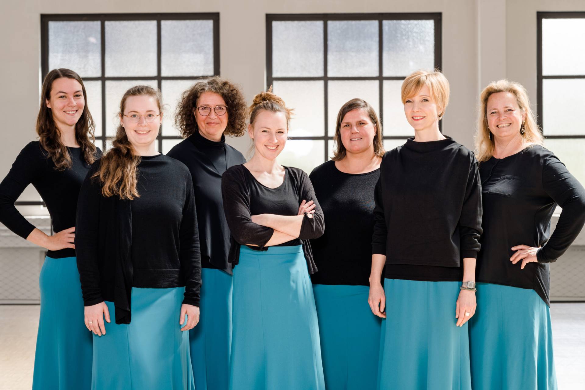 Sieben lächelnde Frauen in blauen Röcken und schwarzen Oberteilen vor einer hellen Fensterfront